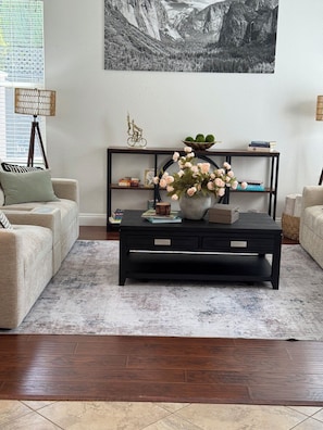 Una sala de estar con una mesa de centro negra, dos sofás color beige, una estantería y un cuadro de un paisaje montañoso en la pared.