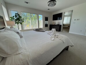 A hotel room with a large bed, a flat-screen TV, a plant, and a view of the outdoors through the windows.