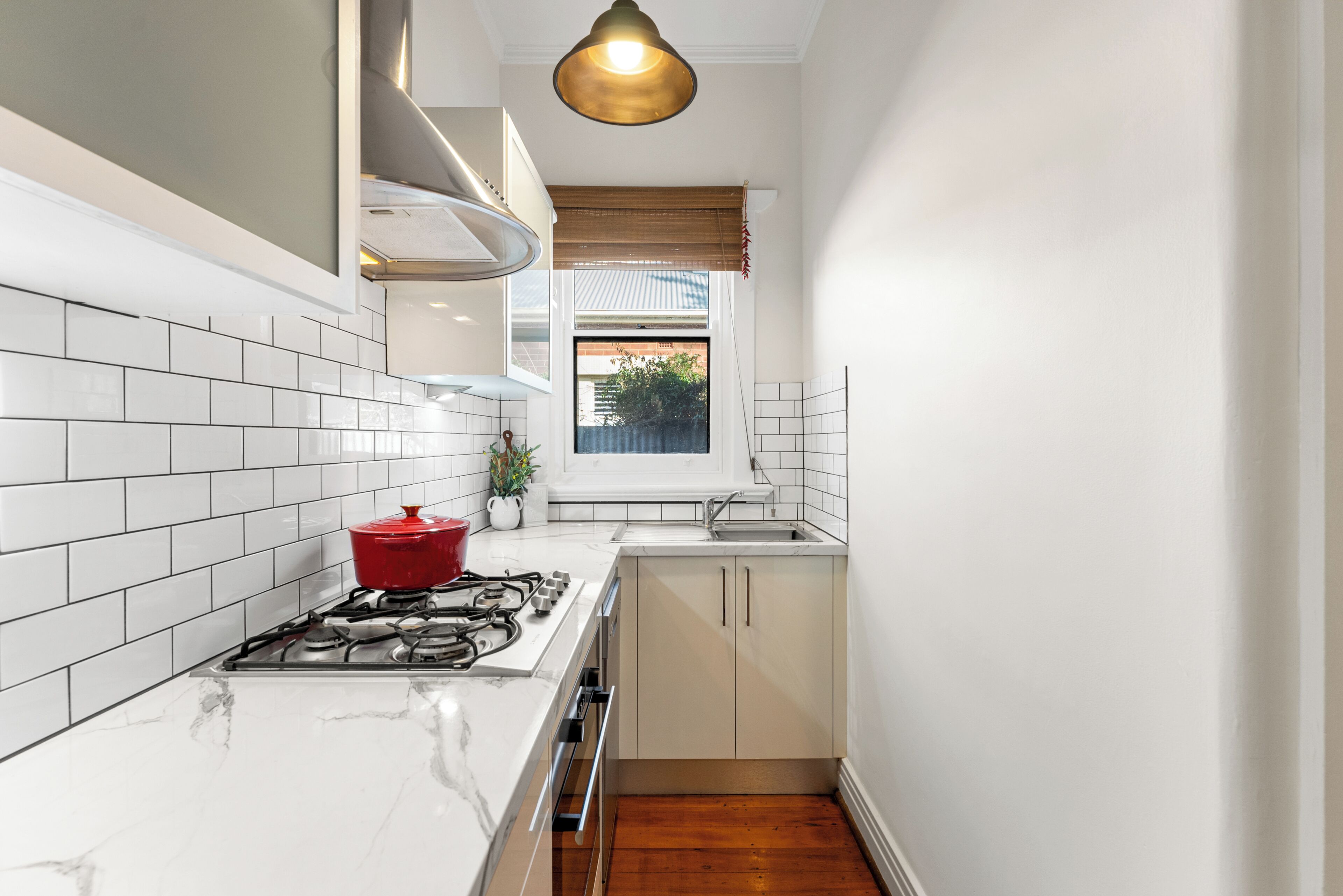 A compact kitchen with white tiles, a gas stove, a red pot, and a window with a view of greenery.