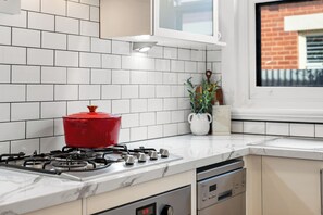 A kitchen with white tiled walls, a red pot on the stove, a window with a view of a building, and a potted plant on the counter. - Glandore Charm Contemporary Haven near CBD and Coast (Glandore)