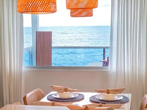 A dining table with two plates and napkins, a view of the sea through a window, and orange hanging lamps.