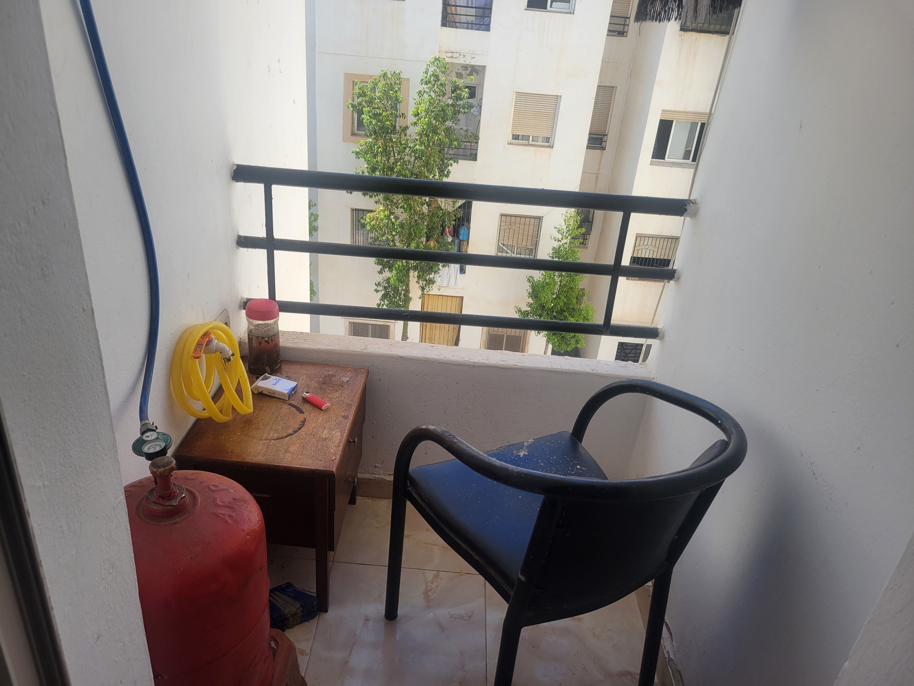Un balcon avec une chaise, une table, une bouteille de gaz rouge et un tuyau jaune.