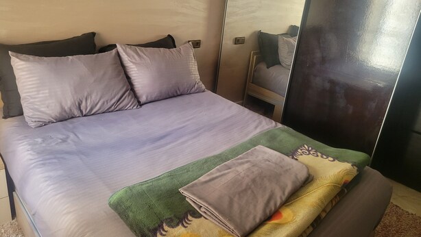 Een bed met een grijze matras en kussens, een groene dekbedovertrek en een opgevouwen grijze deken op een bruine bank.