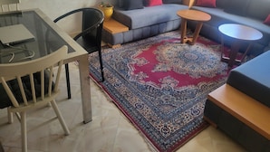 Een woonkamer met een glazen tafel, een bank met kussens, een kamerkleed met dessin en een kleine ronde tafel.