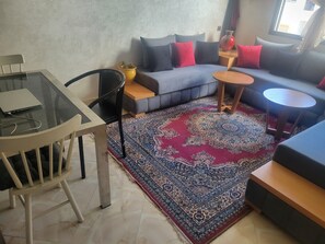A living room with a glass table, a sofa with cushions, a patterned rug, and a small round table.