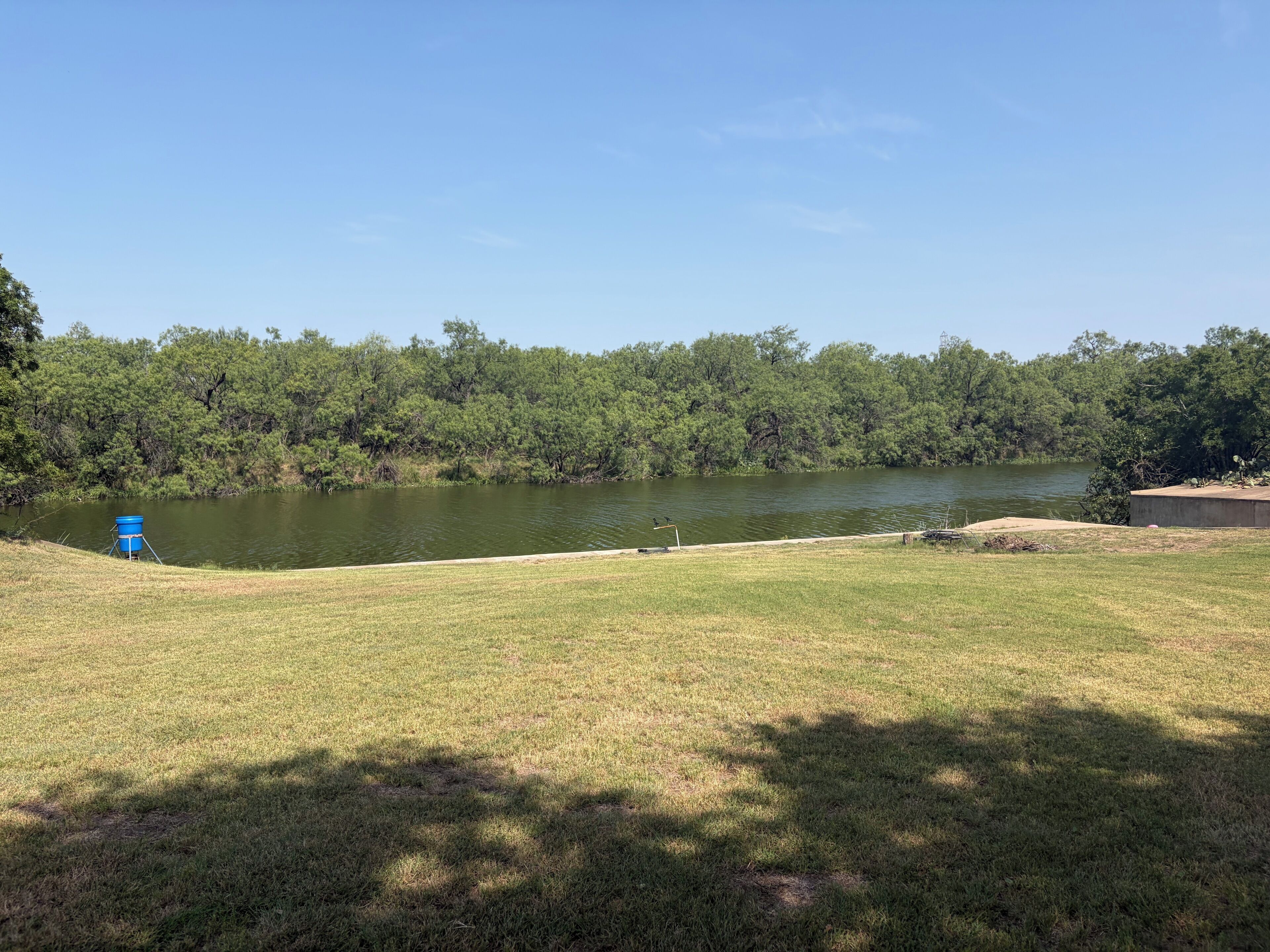 A grassy area with a body of water, trees, and a blue container.