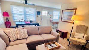 A living room with a sectional sofa, a small ottoman, a chair, a dining table with chairs, and a lamp.