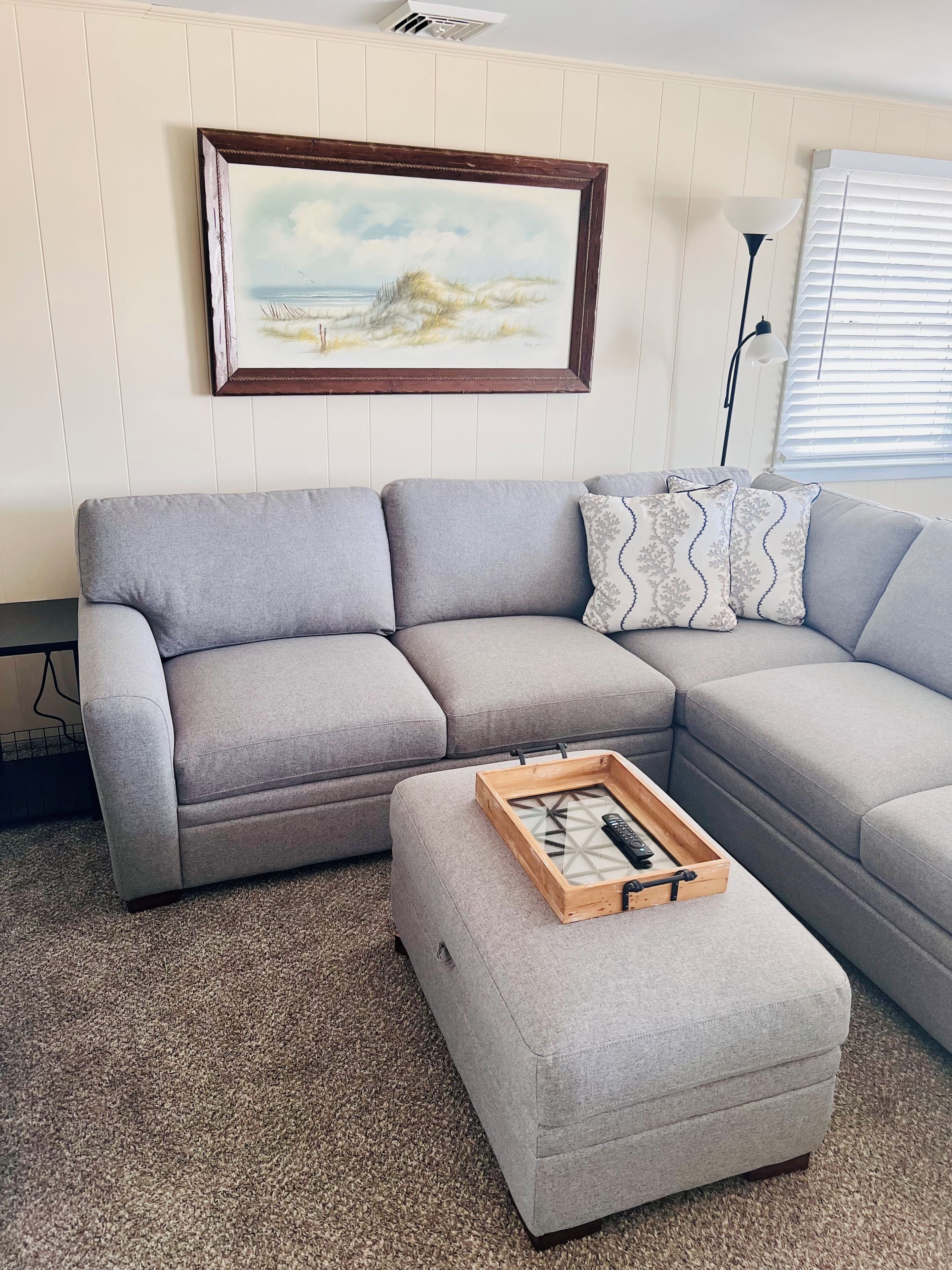 A living room with a grey sectional sofa, a small ottoman, a framed landscape painting, a floor lamp, and a window with blinds.