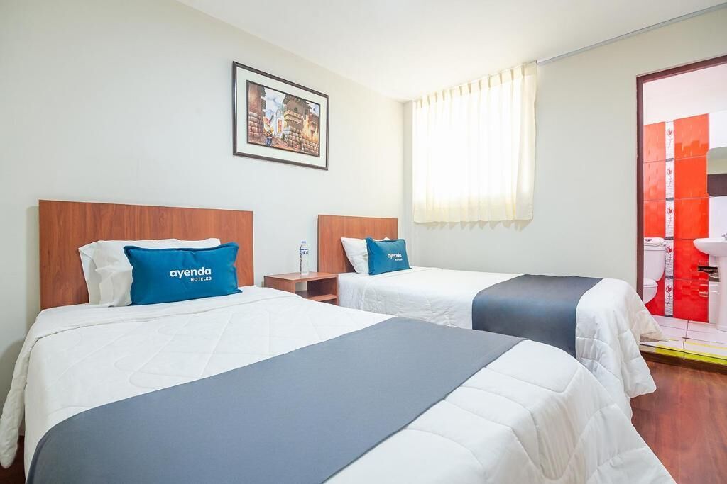 A hotel room with two beds, each with a pillow labeled 'ayenda hotel.' There is a framed picture on the wall and a window with curtains.