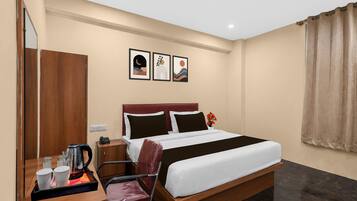 Quarto de hotel com uma cama, escrivaninha com chaleira, chávenas e uma cadeira. Três quadros emoldurados na parede.