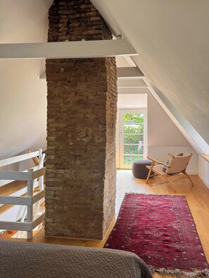 A loft bedroom with exposed brick, a white railing, a red patterned rug, and a view of greenery outside. - Historic house from 1827 on Langeland (Rudkøbing)