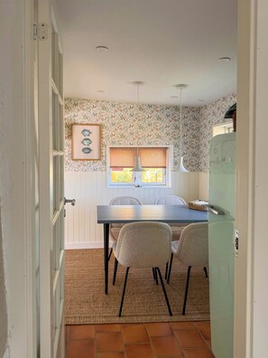 A dining area with a table, chairs, a window with blinds, and a wall with floral wallpaper.