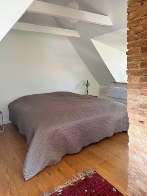 A bedroom with a sloped ceiling, a bed with a patterned bedspread, a small bedside table with a lamp, and a brick wall.