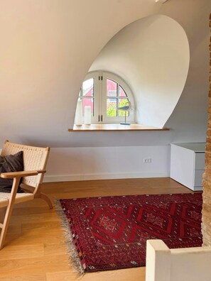 A room with a curved ceiling, a red patterned rug, a wicker chair, and a window with a view of a red building. - Historic house from 1827 on Langeland (Rudkøbing)