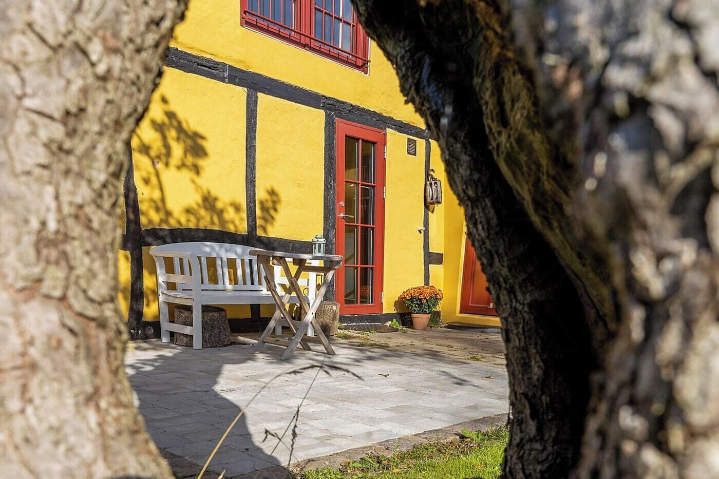 A yellow building with a red door and window, a white bench, and a table with a lantern.