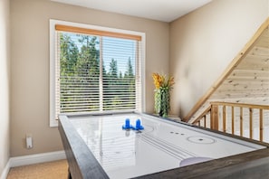 A room with a shuffleboard table, a window with blinds, a plant, and a wooden staircase. - Sunlit Mountain Retreat Near Lakes & Ski Fun (Arnold)