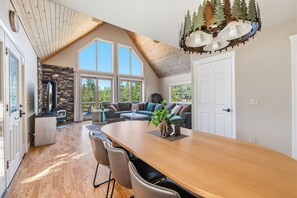 A spacious living area with a large wooden dining table, a stone fireplace, and a view of the outdoors through large windows. - Sunlit Mountain Retreat Near Lakes & Ski Fun (Arnold)