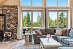 A cozy living room with a stone fireplace, a sofa set, a coffee table, and large windows offering a view of trees. - Sunlit Mountain Retreat Near Lakes & Ski Fun (Arnold)