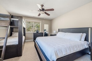 Ein Schlafzimmer mit Etagenbett, Einzelbett, Deckenventilator, Fenster mit Jalousien, Nachttisch, Lampe und Kommode.