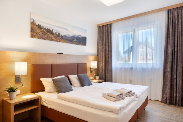 Habitación de hotel con una cama grande, dos mesitas de noche, una lámpara de pared, una planta en maceta y una ventana con cortinas.