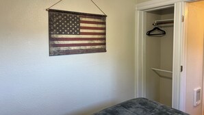 Une pièce dans laquelle on peut voir un drapeau américain suspendu, une armoire avec un cintre, et un banc recouvert d’un tissu gris.