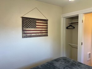 A room with a hanging American flag, a closet with a hanger, and a gray upholstered bench.