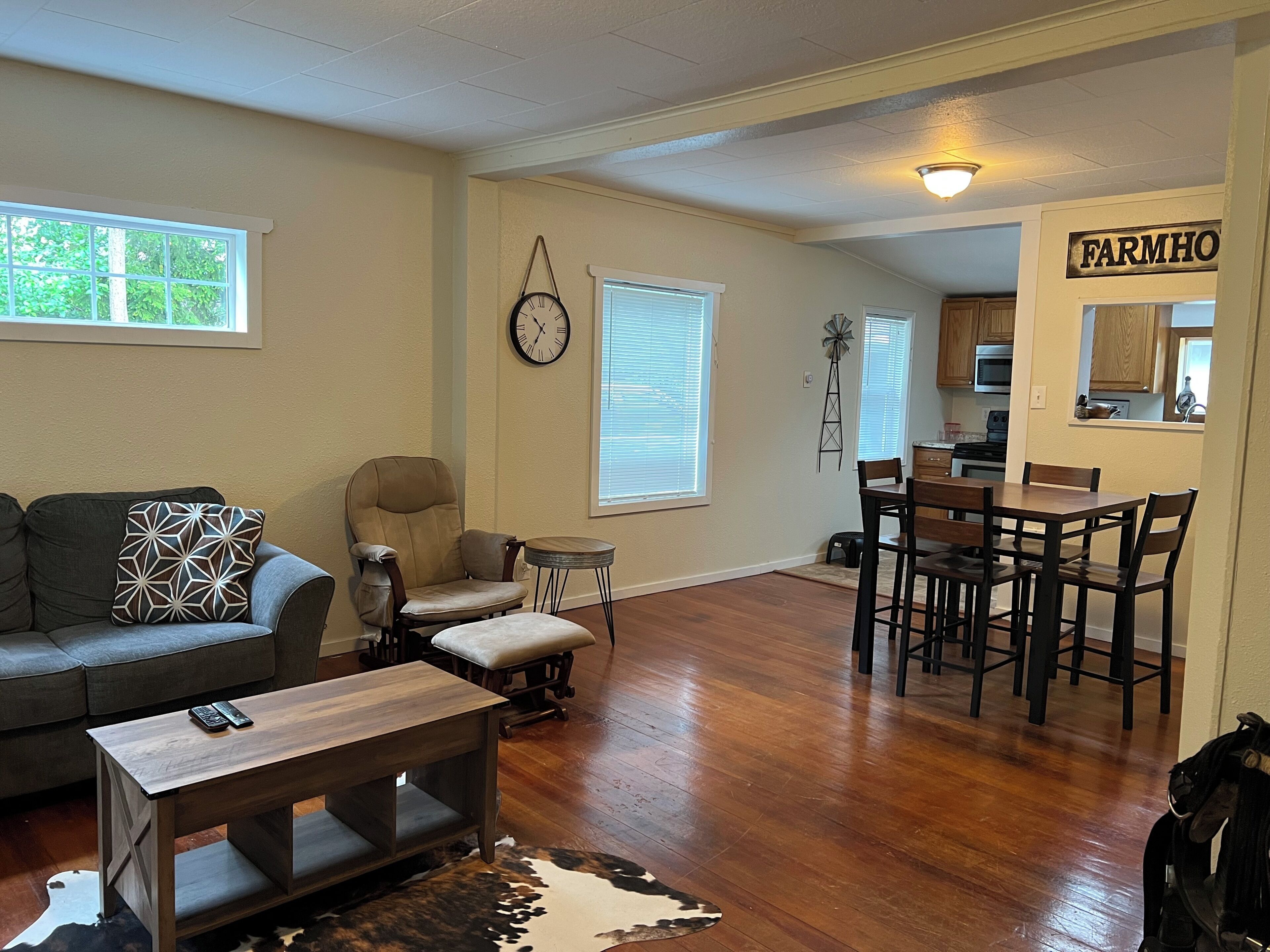 A living room with a sofa, a coffee table, a dining table with chairs, a clock on the wall, and a kitchen area with a sign that reads 'FARMHO'.