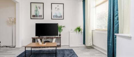 A modern living room with a flat-screen TV, two framed artworks, a coffee table, and a potted plant.
