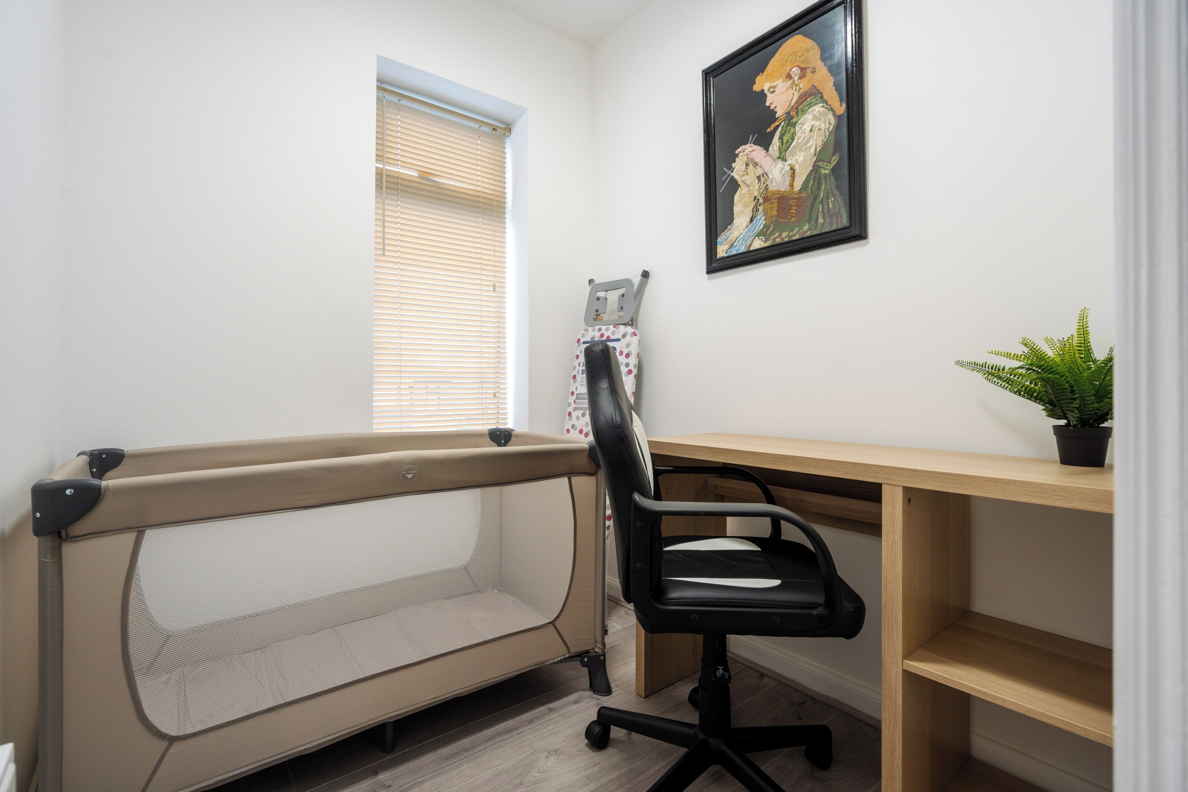 Een kamer met een bed, een bureau, een stoel, een raam met zonwering, een schilderij en een potplant.