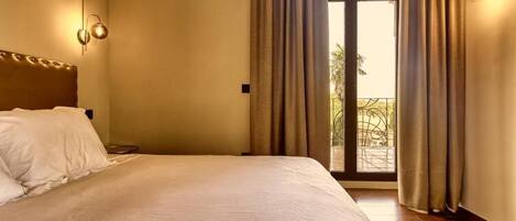 Habitación de hotel con una cama grande, una mesita de noche, una lámpara de pared y vistas al exterior a través de una puerta.