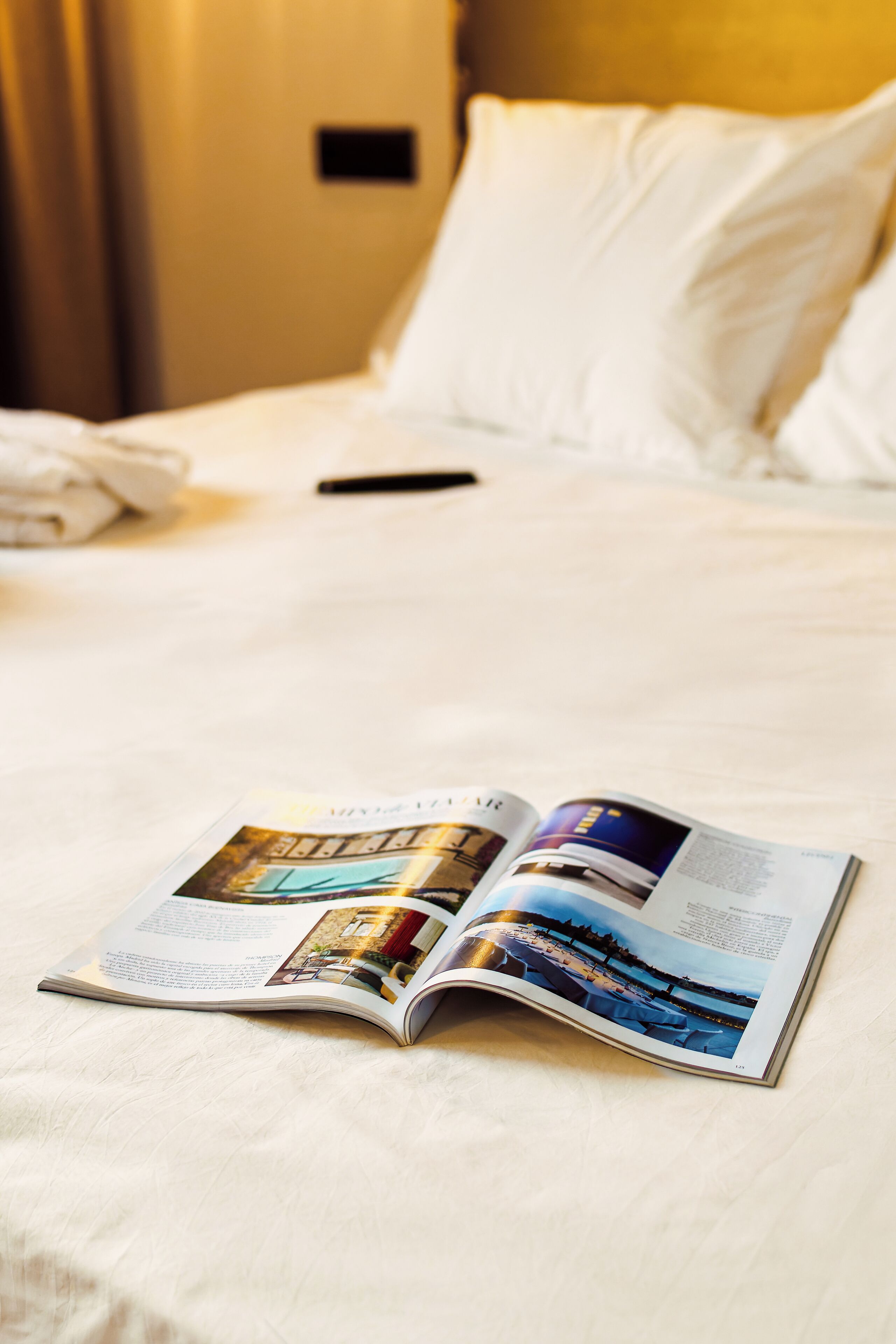 A hotel room with a bed, a pillow, a remote control, and a magazine on the bed.