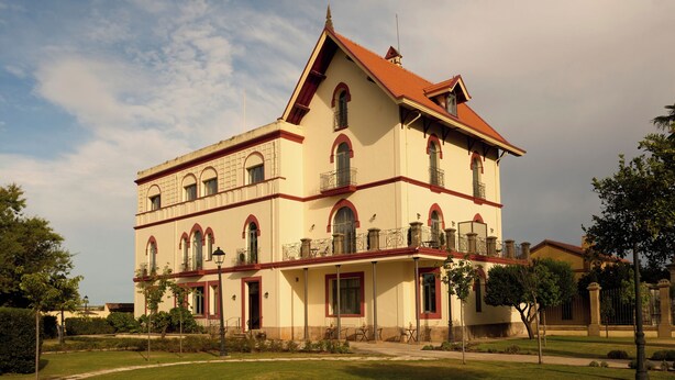 Un edificio de dos pisos con techo de tejas rojas, varios ventanales con balcones y un jardín delantero.