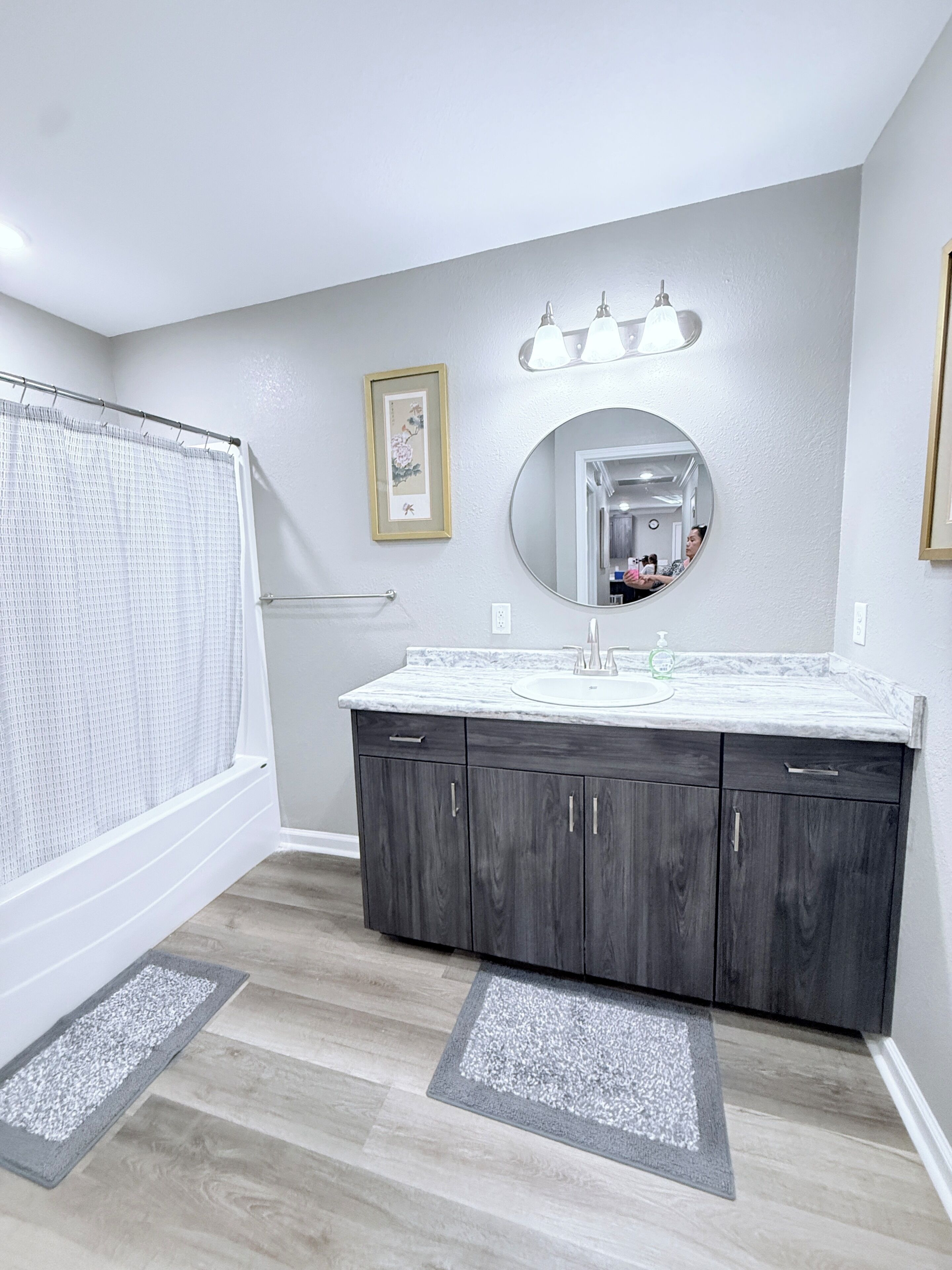A bathroom with a shower, a sink with a marble countertop, a large mirror, and a framed picture on the wall.