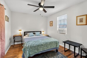 Ein Schlafzimmer mit einem Bett, zwei Nachttischen, einem Deckenventilator, einem Fenster mit Jalousien und einer Wand-Klimaanlage.