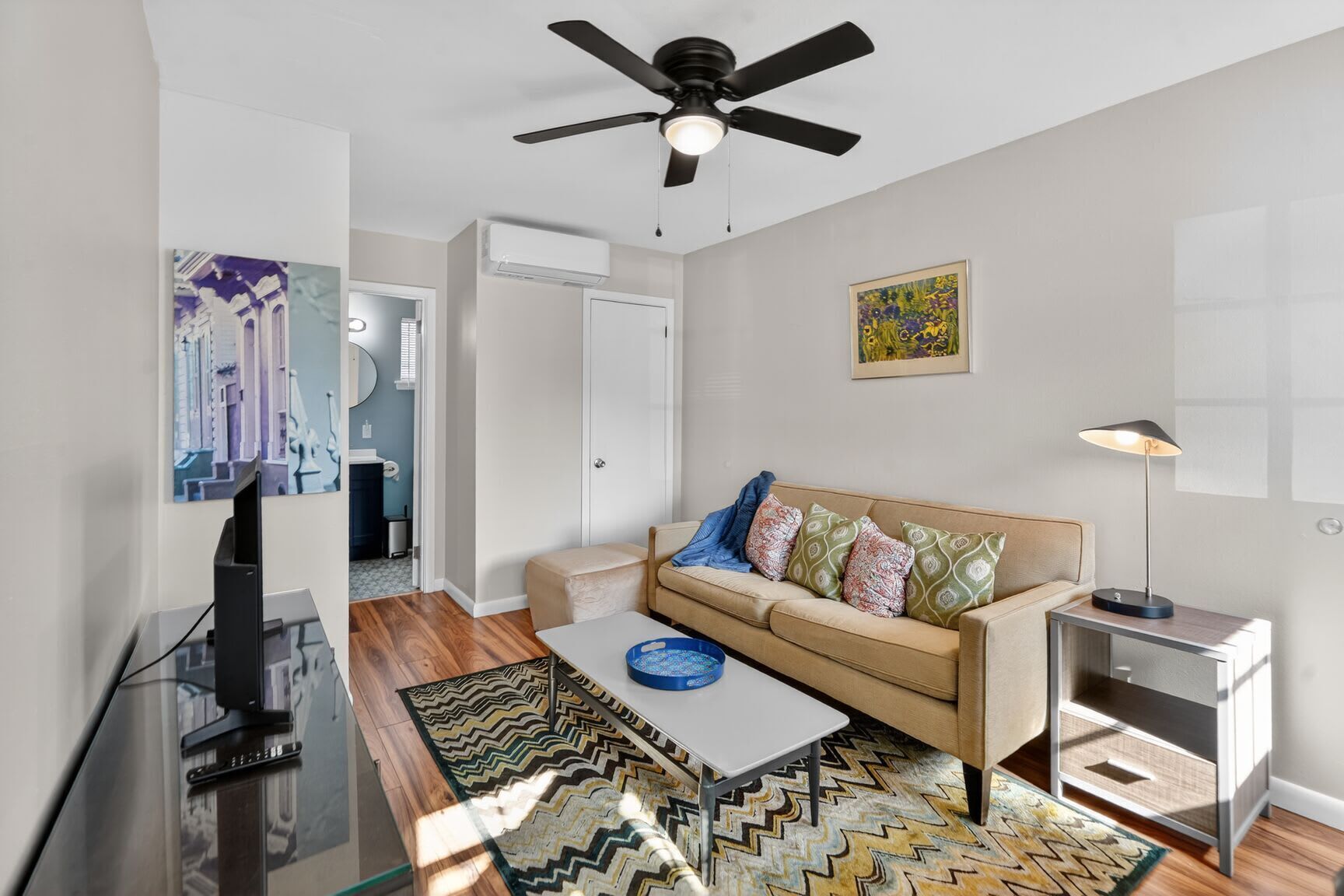 A modern living room with a sofa, coffee table, and a TV. There is a ceiling fan, a painting on the wall, and a lamp on a side table.