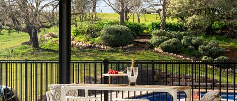 A covered outdoor seating area with a pool, white chairs, and a table with a bottle and glasses.