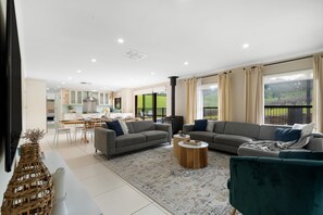 A modern living room with a sofa, armchairs, a coffee table, and a view of the kitchen and outdoor area. - Whispering Vines Scenic Country Retreat with Pool (Balhannah)