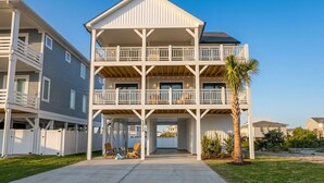 A multi-story residential building with a balcony on each floor, a covered entryway, and a palm tree in the foreground. - Sweet SeaRenity- SURF CITY -New Luxury 4B, Ocean Views, Steps to Beach, Elevator (Surf City)