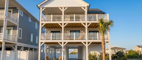 A multi-story residential building with a balcony on each floor, a covered entryway, and a palm tree in the foreground.