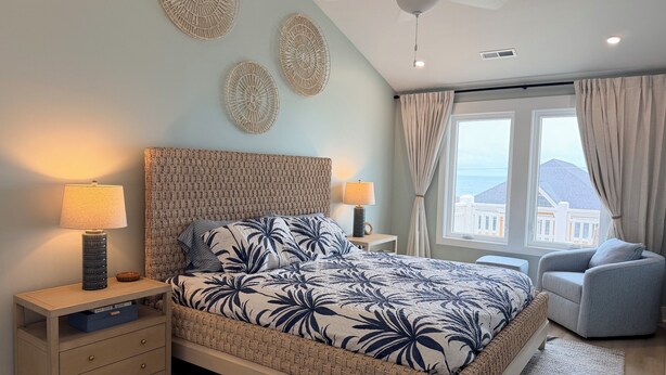 A bedroom with a bed, bedside lamps, a nightstand, a chair, a window with curtains, and a ceiling fan. - Sweet SeaRenity- SURF CITY -New Luxury 4B, Ocean Views, Steps to Beach, Elevator (Surf City)