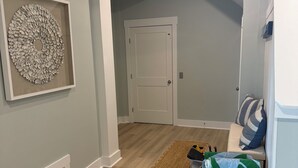 A hallway with a wooden floor, a door, a framed wall decoration, a bench with pillows, and a wicker basket. - Sweet SeaRenity- SURF CITY -New Luxury 4B, Ocean Views, Steps to Beach, Elevator (Surf City)