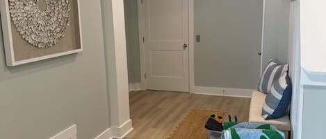Um corredor com piso de madeira, uma porta, um quadro na parede, um banco com almofadas e uma cesta de vime.