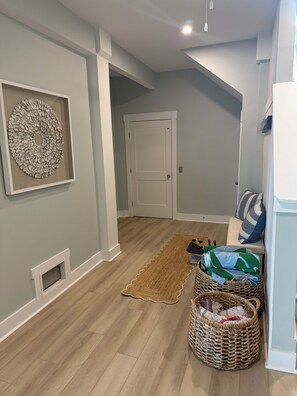 A hallway with a wooden floor, a door, a framed wall decoration, a bench with pillows, and a wicker basket. - Sweet SeaRenity- SURF CITY -New Luxury 4B, Ocean Views, Steps to Beach, Elevator (Surf City)