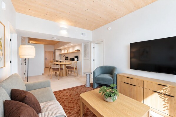 A modern living room with a sofa, a coffee table, and a TV. There is a dining area with a table and chairs, and a kitchen in the background.