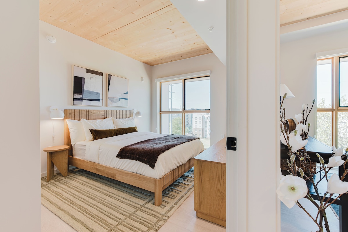 A bedroom with a wooden bed, a bedside table, a lamp, a window, and a view of buildings outside.