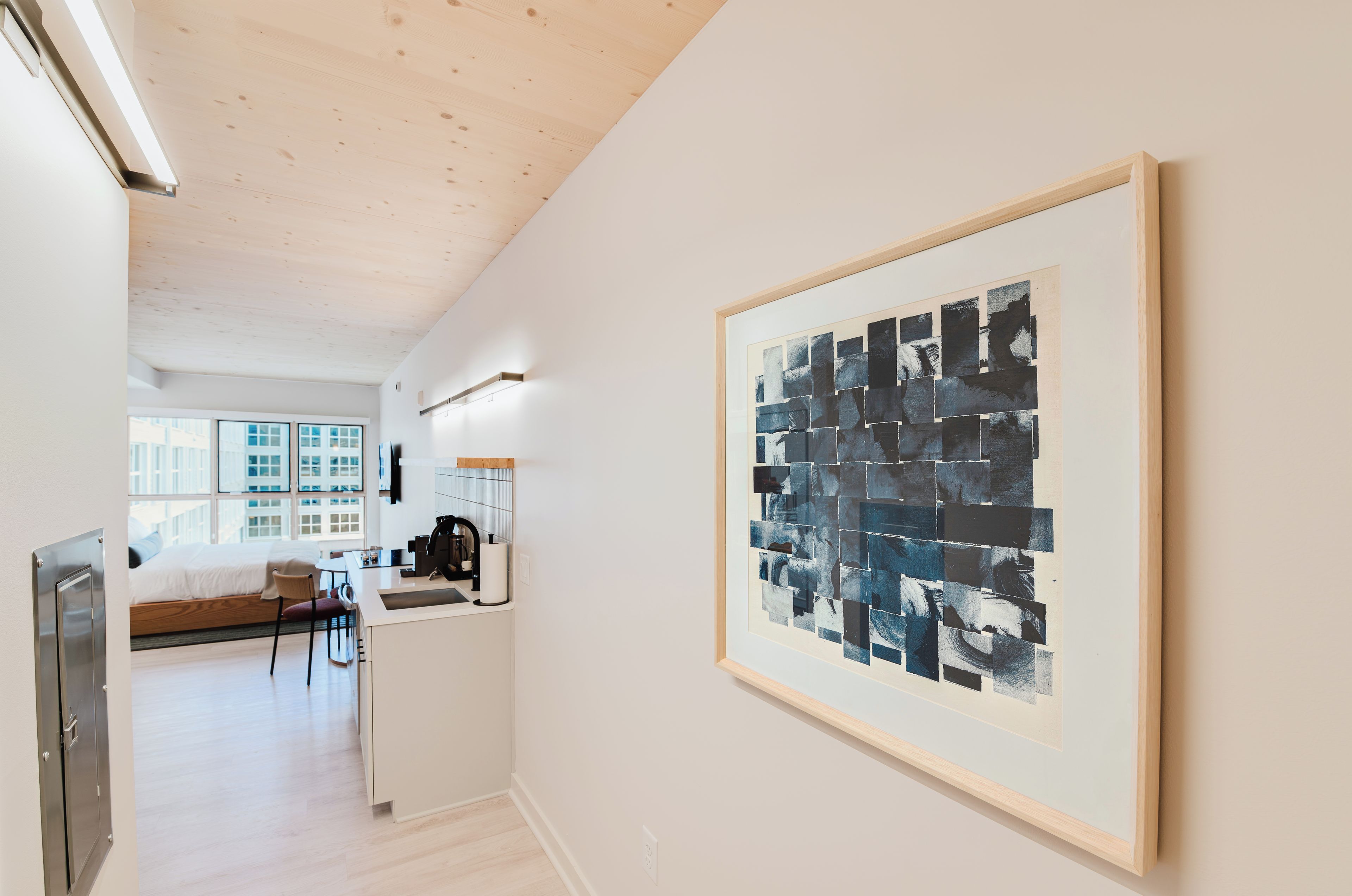 A modern hotel room with a large abstract painting, a kitchenette, and a view of the city through large windows.