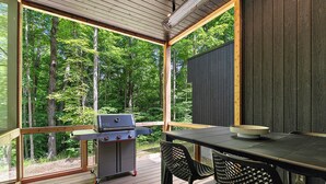 An outdoor deck with a grill, a table, and chairs, surrounded by a forest. - The Linehouse at Norstead (Cedar)