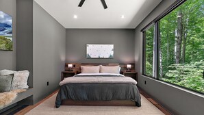 A bedroom with a large bed, a nightstand, a wall-mounted lamp, and a window with a forest view. - The Linehouse at Norstead (Cedar)