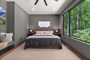 Ein Schlafzimmer mit einem großen Bett, einem Nachttisch, einer Wandlampe und einem Fenster mit Blick auf einen Wald.