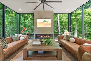 Uma sala de estar moderna com lareira, sofás de couro e uma grande janela com vista para uma floresta.